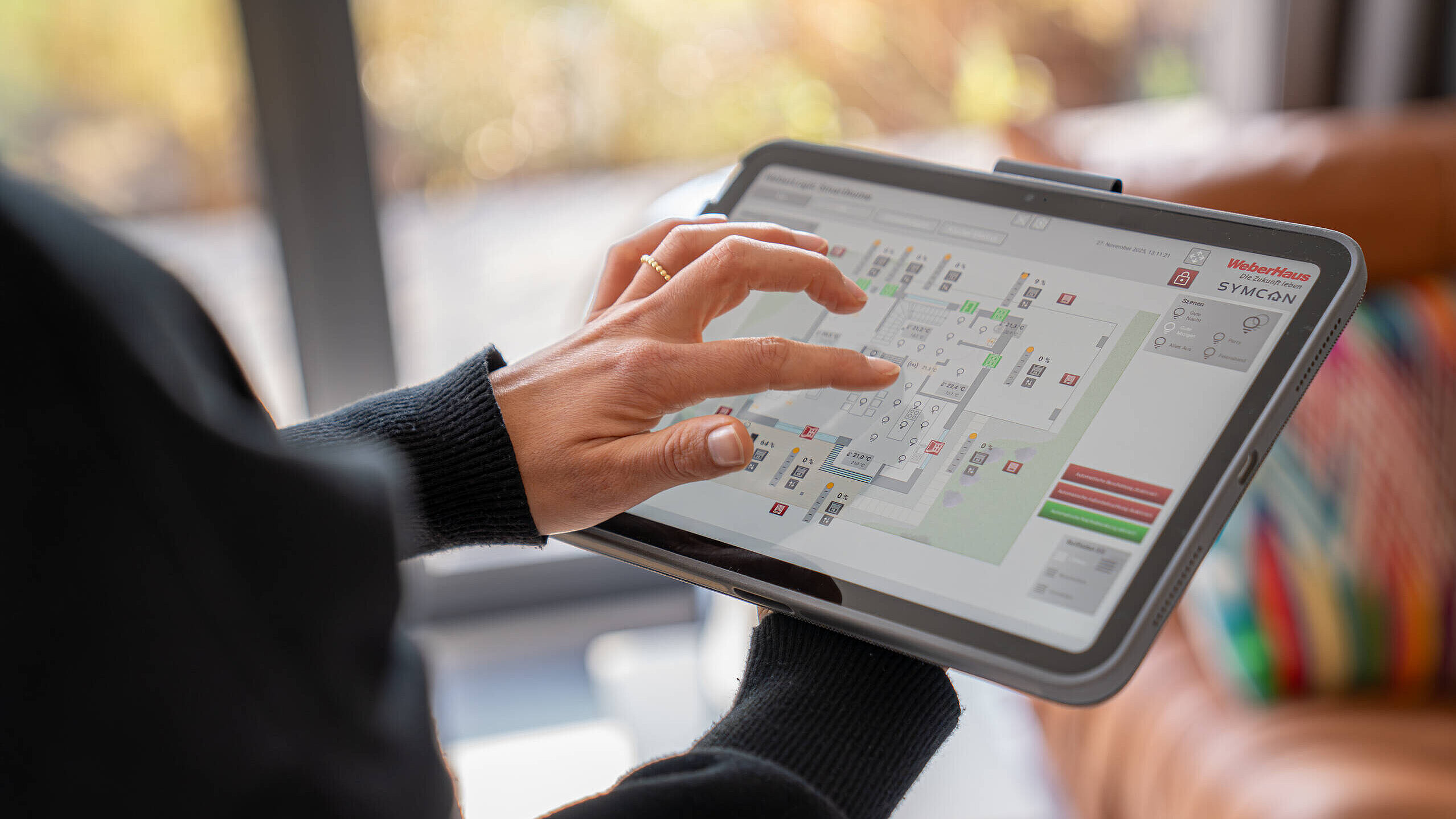 Eine Person nutzt ein Tablet, um einen digitalen Grundriss zu betrachten. Ihre Finger sind auf dem Bildschirm positioniert, während sie eine Interaktion mit der Karte vornimmt. Der Hintergrund zeigt unscharfe Möbel und ein Fenster.