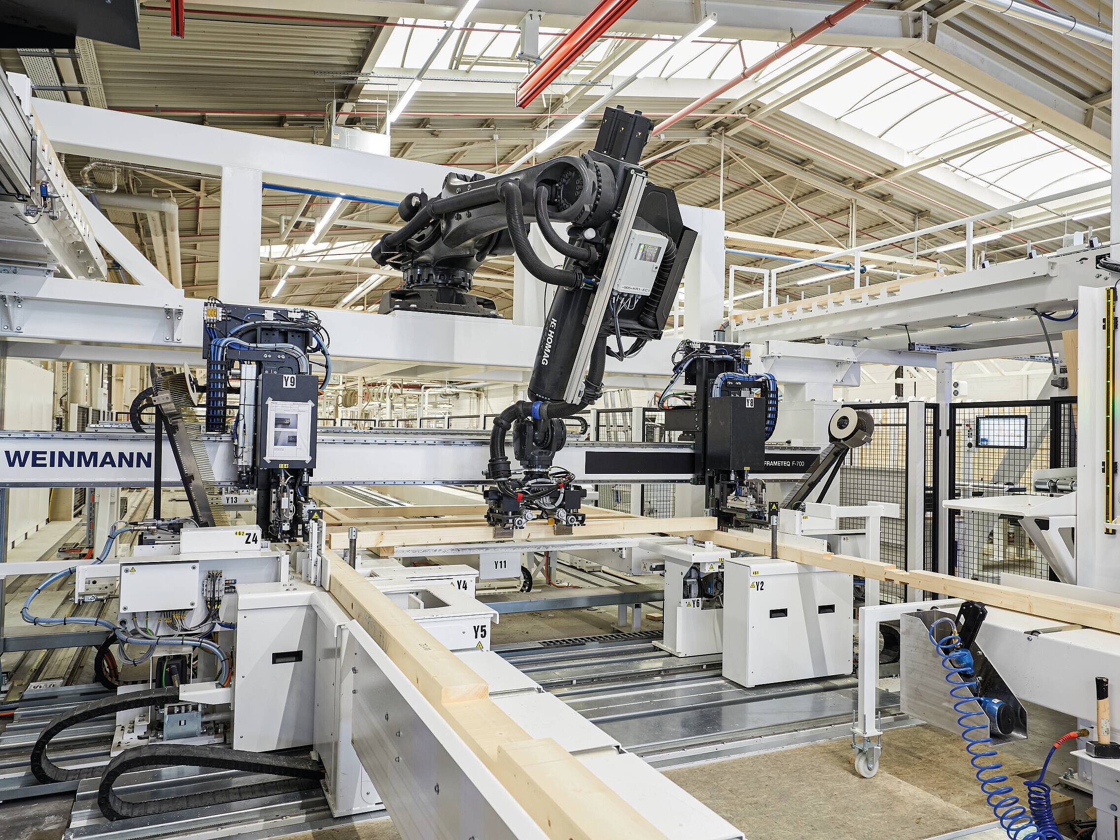 Smarte Robotik im Holzhaus Fertigungsprozess bei WeberHaus Eine moderne automatisierte Maschinenlinie in einer Produktionshalle, die Holzlatten bearbeitet. Roboterarme und Maschinen sind sichtbar, die präzise Arbeiten an den Holzstücken durchführen. Die Umgebung ist hell erleuchtet und technisch ausgestattet.