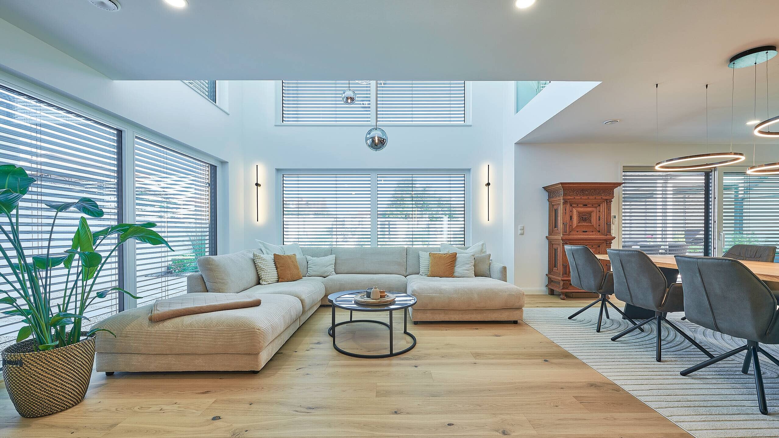 Offenes Raumkonzept Ein modernes Wohnzimmer mit einem großen, beigen Sofa, einem runden Couchtisch und einem dekorativen Pflanzenarrangement. Große Fenster mit Jalousien sorgen für helle Lichtverhältnisse. Im Hintergrund steht ein aus Holz gefertigter Schrank und ein Esstisch mit grauen Stühlen. Der Boden ist aus hellem Holz.