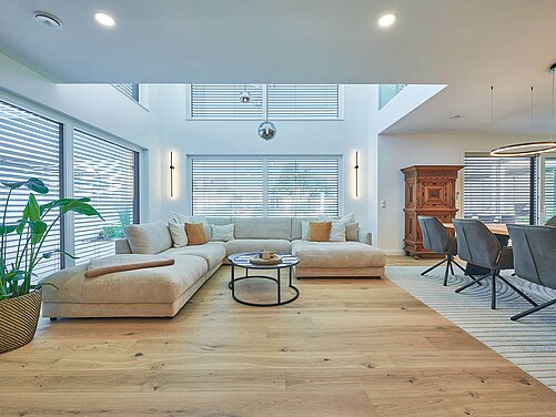 Ein modernes Wohnzimmer mit einem großen, beigen Sofa, einem runden Couchtisch und einem dekorativen Pflanzenarrangement. Große Fenster mit Jalousien sorgen für helle Lichtverhältnisse. Im Hintergrund steht ein aus Holz gefertigter Schrank und ein Esstisch mit grauen Stühlen. Der Boden ist aus hellem Holz.