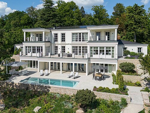 Eine moderne, mehrstöckige Villa mit großen Fenstern und Balkonen, umgeben von üppigem Grün und Bäumen. Vor dem Haus befindet sich ein Pool, auf der Terrasse stehen Tisch und Stühle. Der Garten ist farbenfroh gestaltet, mit verschiedenen Pflanzen und einer gepflasterten Auffahrt.