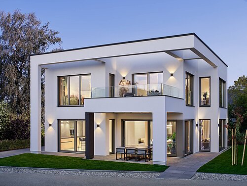 Ein modernes, zweigeschossiges Wohnhaus mit flachem Dach. Es hat große Fenster, die Licht hereinlassen, und einen Balkon. Die Fassade ist hell und schlicht, umgeben von einem gepflegten Garten mit Rasen und wenigen Pflanzen. Der Himmel ist leicht blau, was eine angenehme Abendstimmung vermittelt.