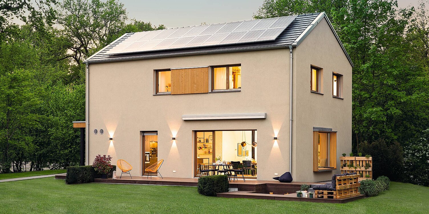 Ausstellungshaus sunshine Ein modernes, zweistöckiges Haus mit Solarplatten auf dem Dach. Die Fassade ist in hellen Farbtönen gehalten und hat große Fenster. Vor dem Haus befindet sich eine grüne Rasenfläche und ein Holzdeck mit Sitzmöglichkeiten. Umgeben von Bäumen und Sträuchern.