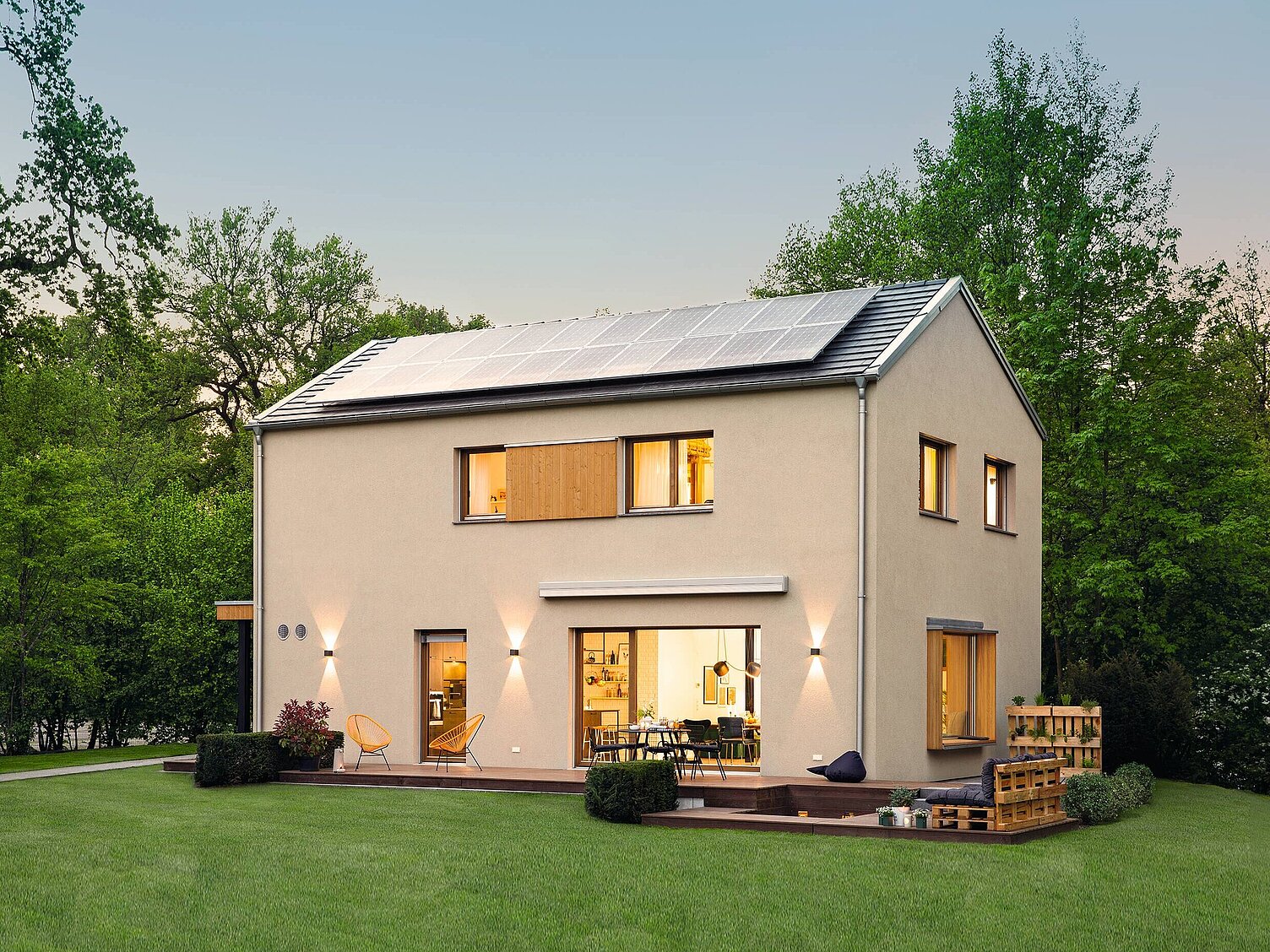 Ein modernes, zweistöckiges Haus mit Solarplatten auf dem Dach. Die Fassade ist in hellen Farbtönen gehalten und hat große Fenster. Vor dem Haus befindet sich eine grüne Rasenfläche und ein Holzdeck mit Sitzmöglichkeiten. Umgeben von Bäumen und Sträuchern.