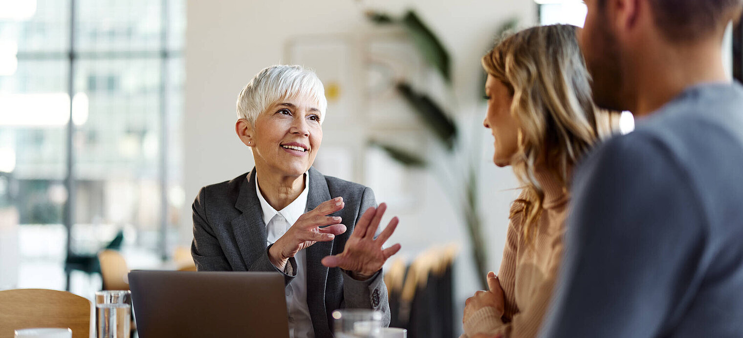 Eine Beraterin mit kurzem grauen Haar und einem freundlichen Lächeln spricht mit einem jungen Paar in einem modernen Büro. Vor ihr steht ein aufgeklappter Laptop, die Atmosphäre wirkt professionell und zugleich entspannt.