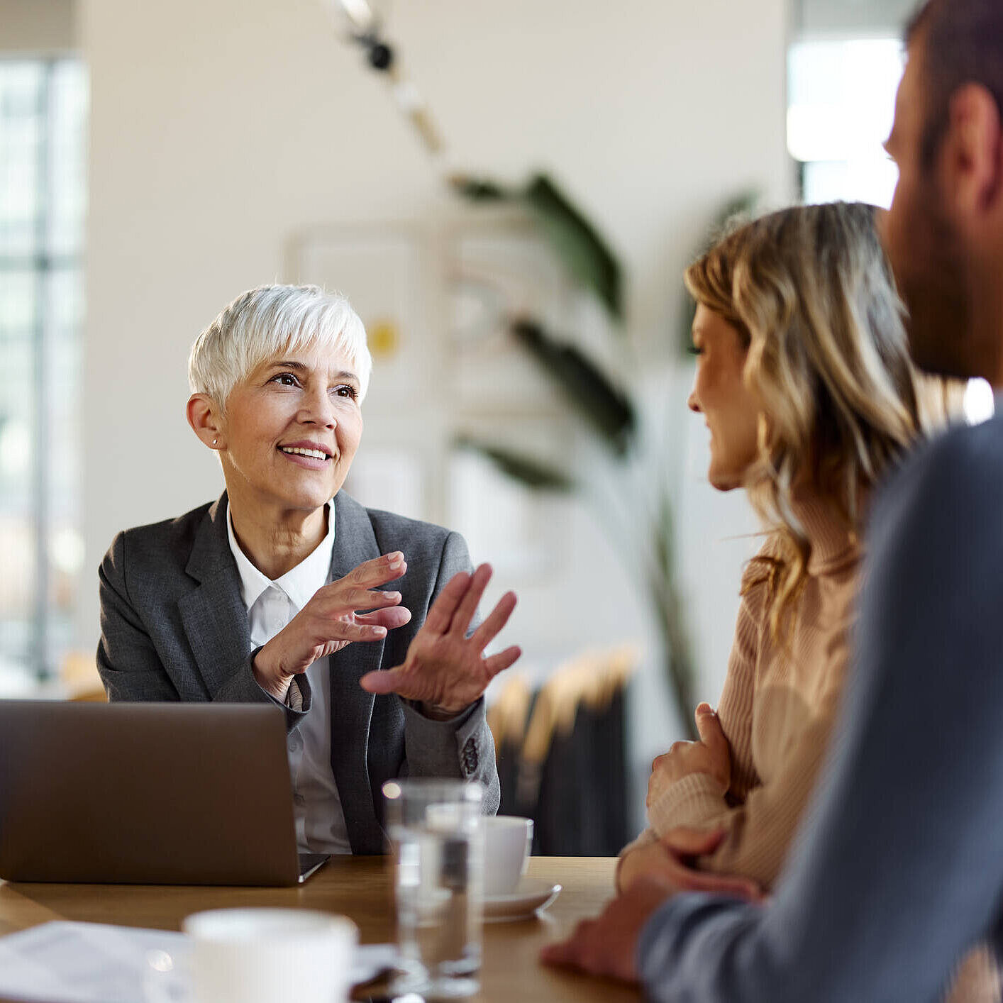 Eine Beraterin mit kurzem grauen Haar und einem freundlichen Lächeln spricht mit einem jungen Paar in einem modernen Büro. Vor ihr steht ein aufgeklappter Laptop, die Atmosphäre wirkt professionell und zugleich entspannt.