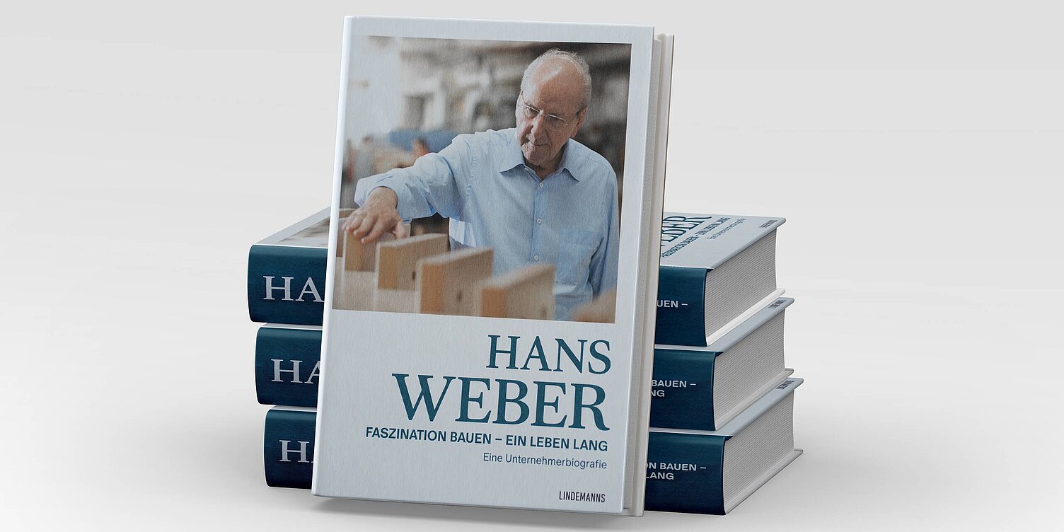 Ein Buch mit dem Titel „Hans Weber: Faszination bauen – ein Leben lang“ steht im Vordergrund. Auf dem Cover ist ein älterer Mann zu sehen, der konzentriert an einem Tisch mit Holzteilen arbeitet. Im Hintergrund sind unscharfe Werkstattutensilien zu erkennen.